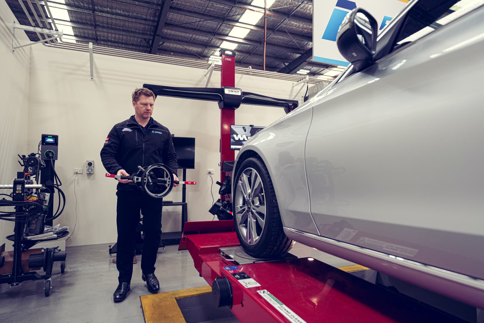Technician carrying a wheel alignment clamp beside an alignment system